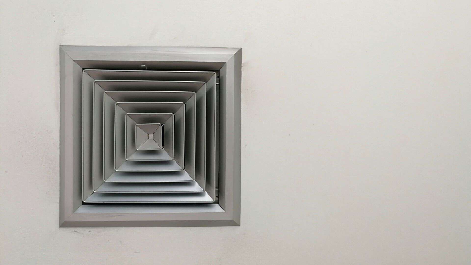 A silver, square air vent with a louvered design is mounted on a plain white wall, resembling the layered precision one might admire after a thorough air duct cleaning. The surrounding wall remains smooth and unadorned.