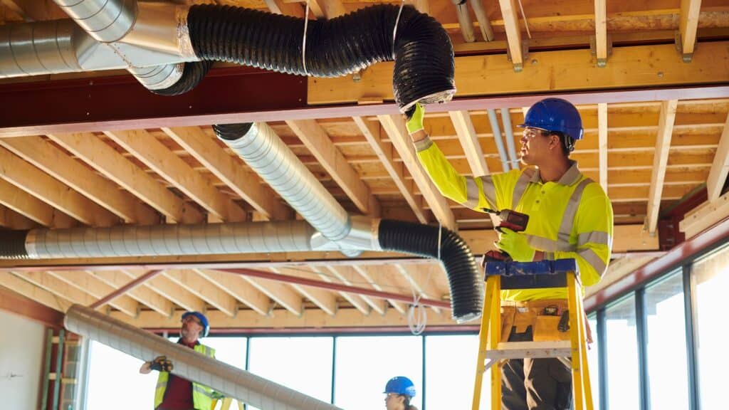 Three construction workers, well-versed in dryer vent cleaning, install ductwork in a building with exposed wooden beams. One stands on a ladder with a power drill, while others support the ventilation pipes. All wear blue hard hats and safety gear for utmost safety.