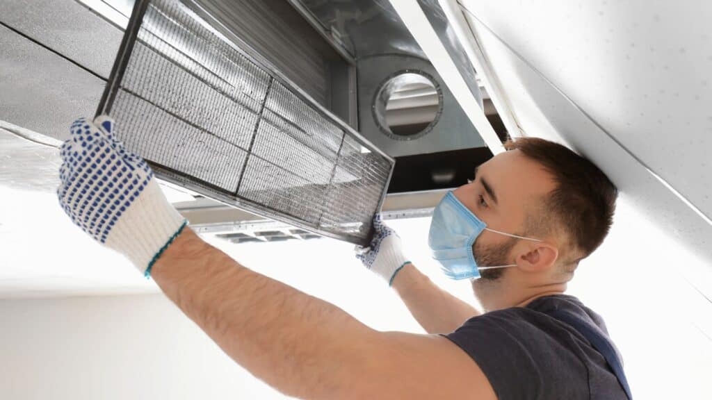 A person wearing a blue mask and gloves is meticulously installing or removing a filter from a ceiling air vent, resembling the precision needed in air duct cleaning. They are looking up and handling the filter with care.