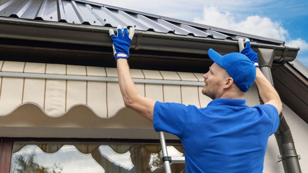 A man in a blue uniform and gloves is standing on a ladder, expertly performing gutter cleaning on the roof of a house. Engaged in maintenance work, he ensures everything flows smoothly, with a striped awning visible beneath the gutter.
