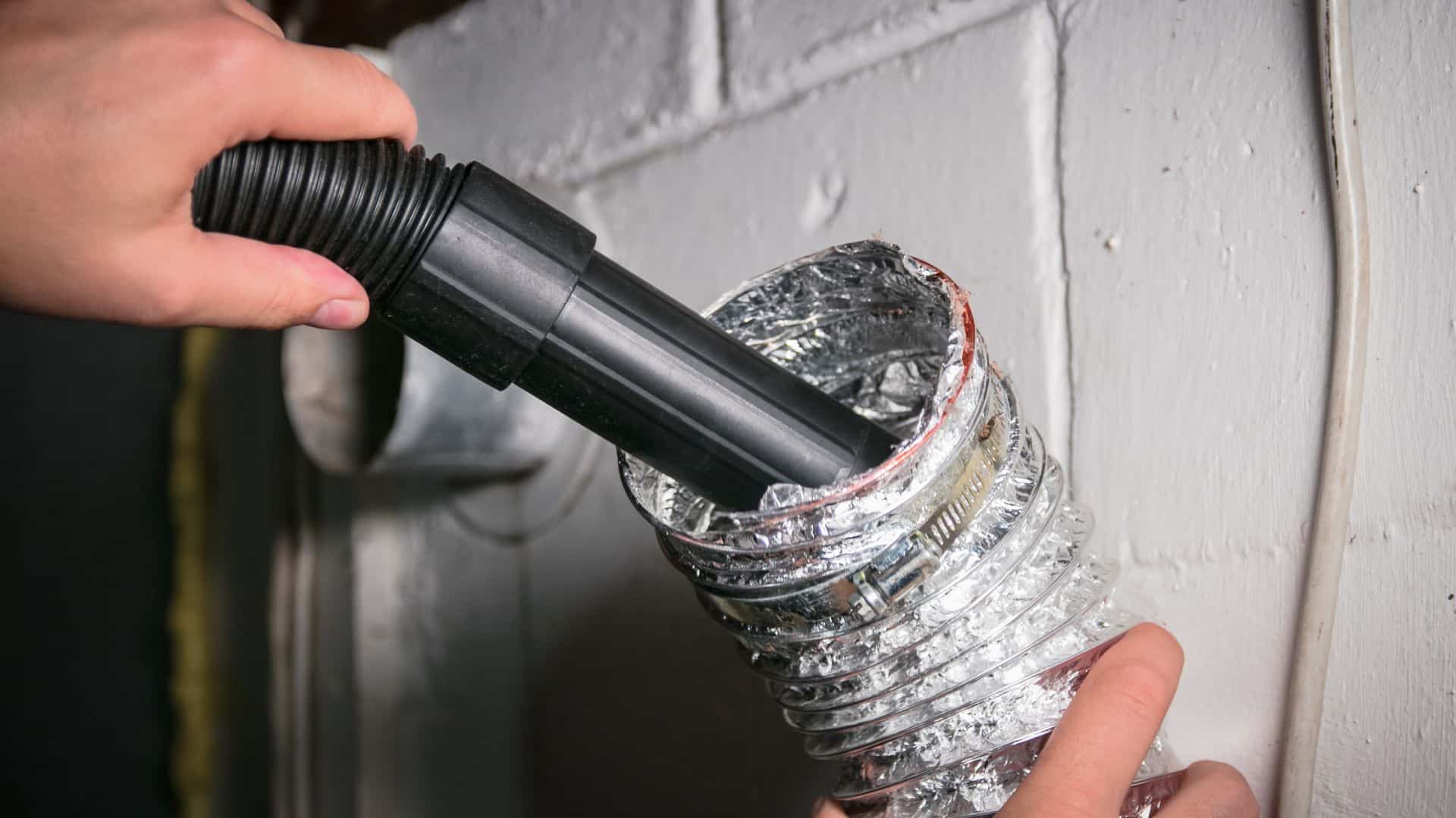 A person uses a vacuum attachment for air duct cleaning inside a flexible metal vent duct against a white brick wall.