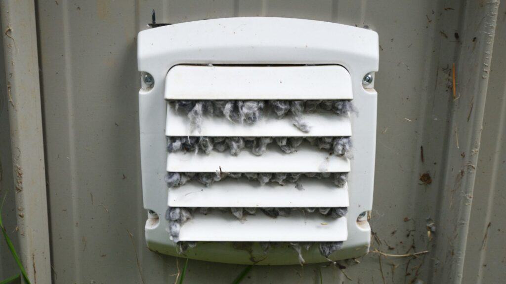 A white vent, mounted on a beige wall, is clogged with gray lint and debris, highlighting the urgent need for air duct cleaning.