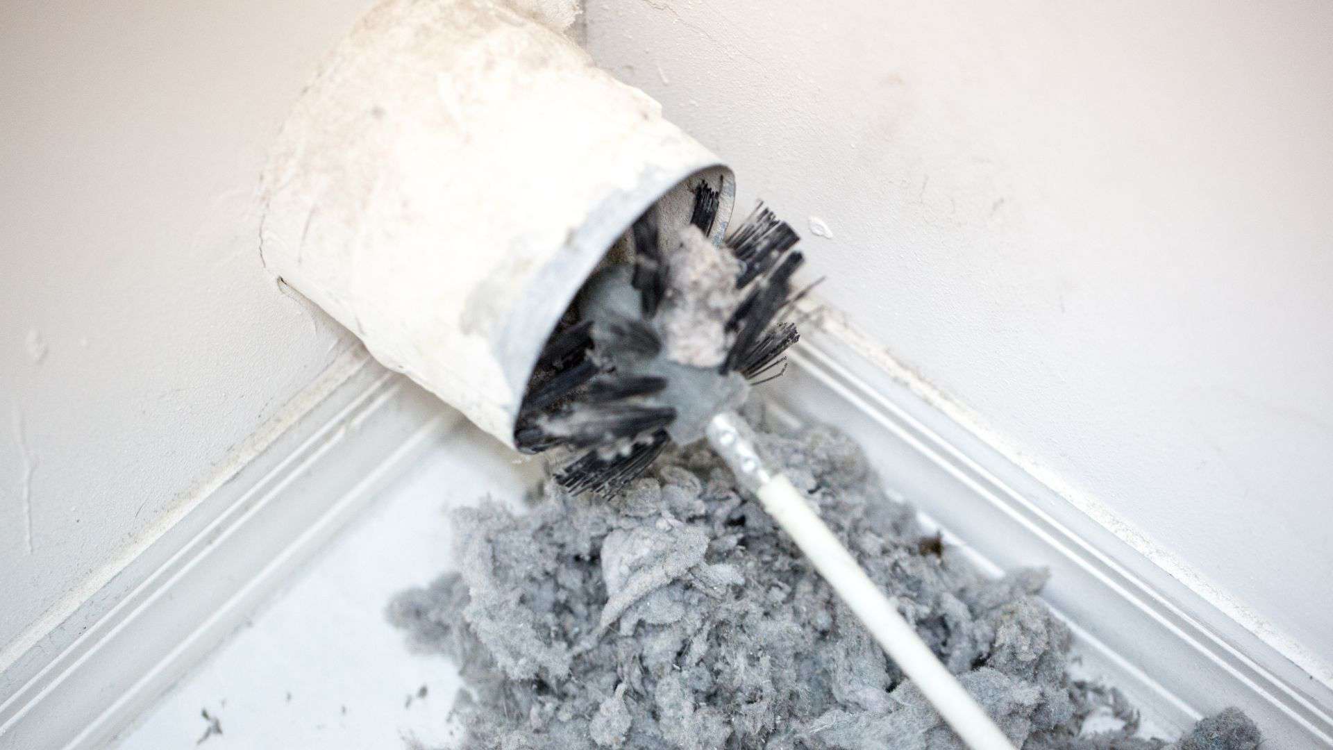 A white toilet brush stands in its holder in the corner of a room. The base and brush are covered in gray dust and lint, resembling the neglect of air duct cleaning, with a pile accumulating on the white floor beneath. The wall is lightly stained.