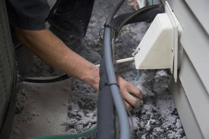 A person reaches into a white external vent on a building, surrounded by debris and connected hoses. The scene suggests maintenance or repair work, possibly involving chimney cleaning. The hand and part of the arm are visible, showing active engagement in the task.