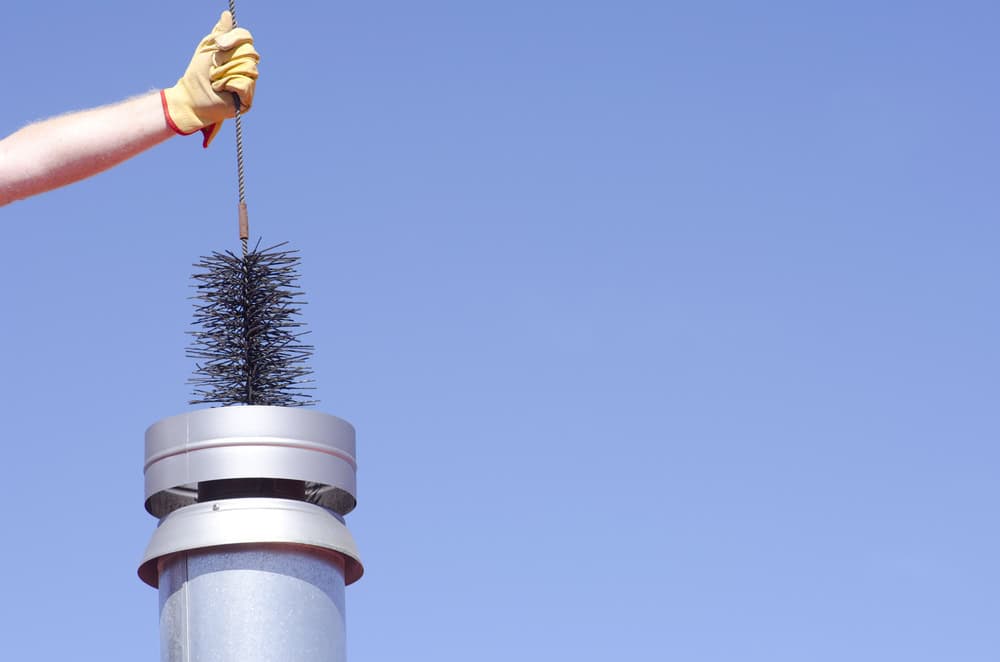 A person wearing a yellow glove is holding a wire chimney brush above the top of a metal chimney pipe against a clear blue sky, showcasing meticulous attention to detail akin to air duct cleaning.