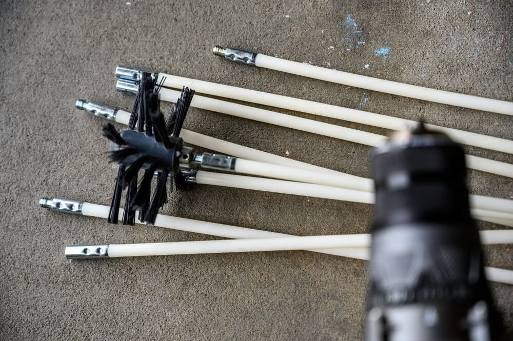 A collection of chimney cleaning rods with metal connectors and a black bristle brush lie on a concrete surface, ready to tackle any chimney cleaning task. The blurred foreground shows a part of a power tool, hinting at its potential use for dryer vent cleaning as well.