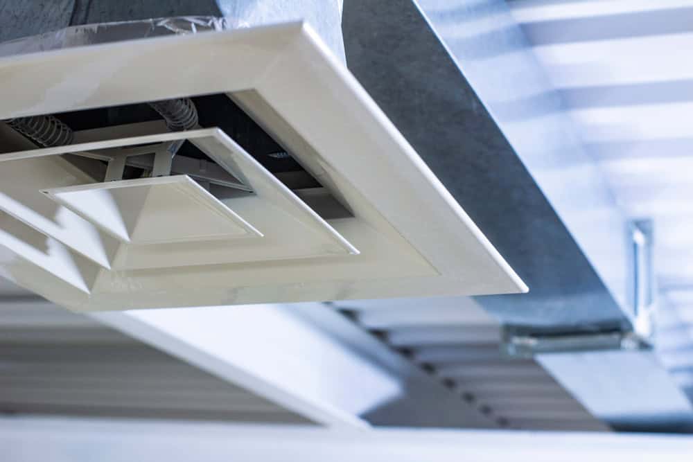 Close-up of a white ceiling air vent with a square, geometric design against a background of exposed metal beams and ductwork in an industrial setting, echoing the meticulous precision found in chimney cleaning.