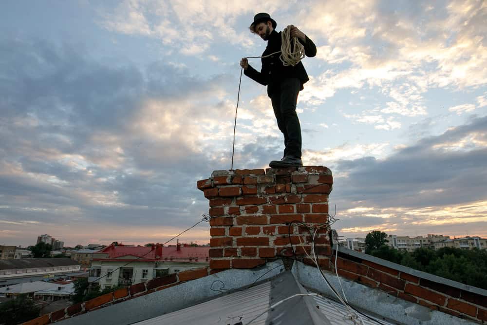 A person stands on a rooftop chimney, ropes coiled for chimney cleaning. The sky is cloudy with hints of blue, and buildings and trees form the backdrop. Clad in dark clothing and a hat, they prepare for their task with precision.