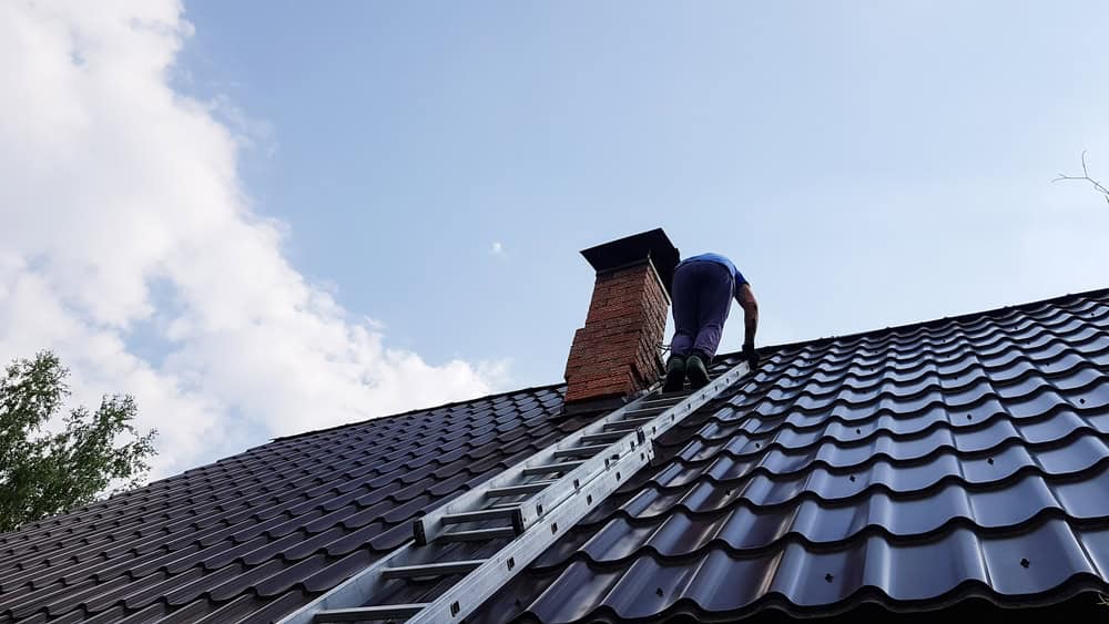 A person climbs a ladder onto a black metal roof next to a brick chimney under a blue sky with clouds, preparing for some thorough gutter cleaning.