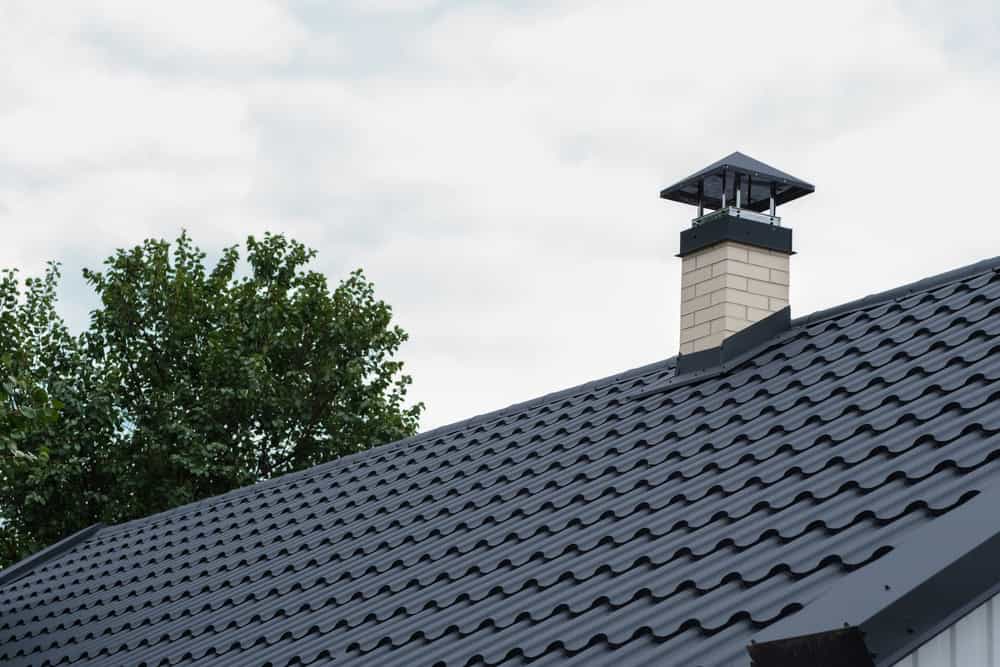 A sloped roof with dark grey metal shingles and a light brick chimney topped with a black cap suggests careful maintenance, such as chimney repair. In the background, a tree with green leaves is partially visible under a cloudy sky.