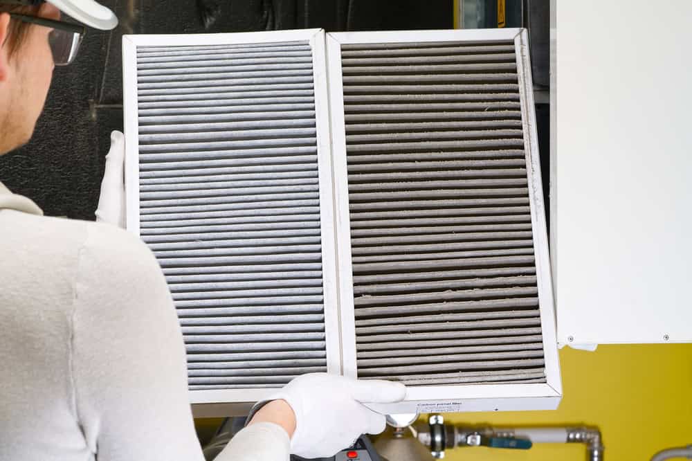 A person wearing gloves holds two air filters, one clean and one dirty, illustrating the contrast in particle buildup. The scene, reminiscent of an air duct cleaning session, is set indoors, possibly in a workshop or maintenance area.