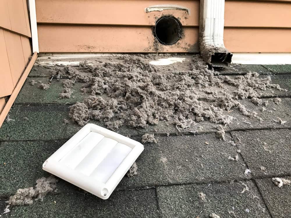 A dryer vent with a removed cover, surrounded by a large amount of accumulated lint on a roof. The exterior wall and part of a gutter are visible, indicating routine maintenance like gutter cleaning or attic insulation is underway.