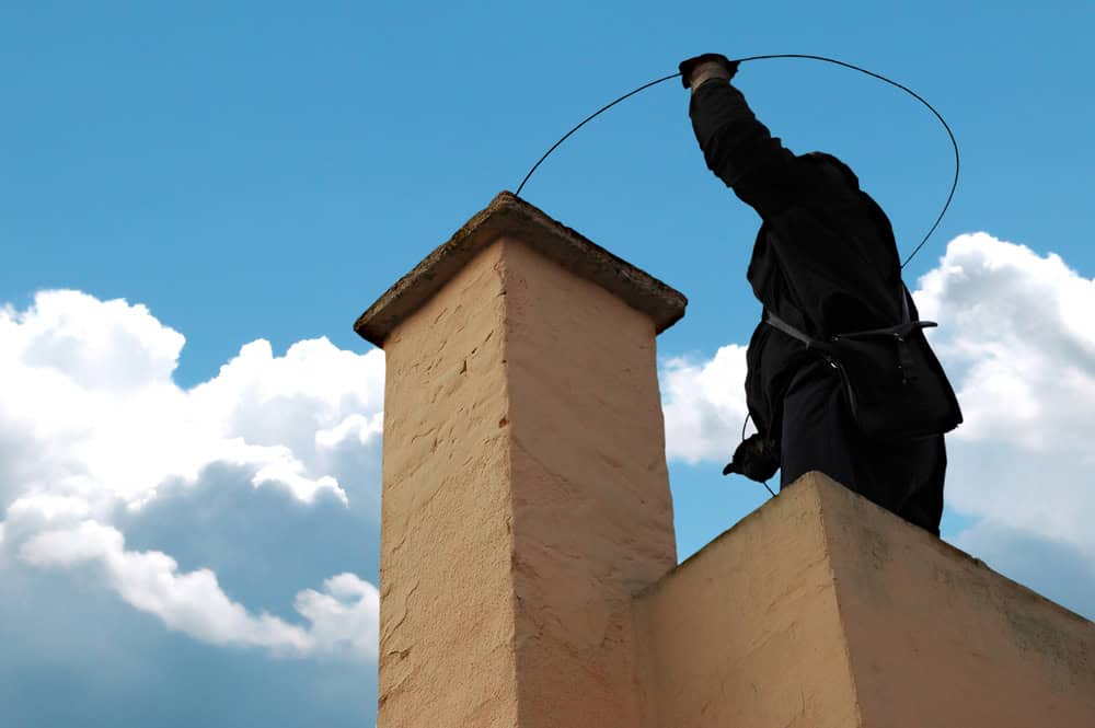 A person in dark clothing expertly performs chimney cleaning on a rooftop, deftly wielding a wire tool against the backdrop of a partly cloudy blue sky.