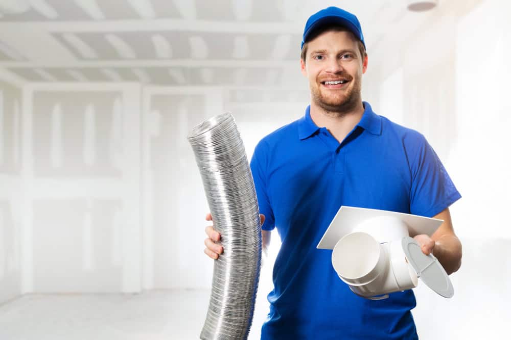 A smiling man in a blue uniform and cap holds an aluminum duct in one hand and a ventilation component in the other, ready for some dryer vent cleaning. He stands in an unfinished room with drywall walls, emphasizing the importance of proper airflow for safety and efficiency.