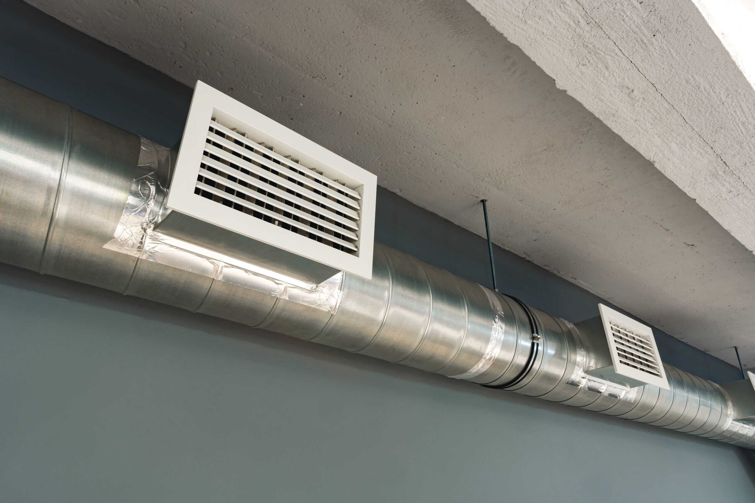 A large metallic air duct runs along a concrete ceiling, featuring two white rectangular vents evenly spaced along its length. The walls are painted in a muted blue-gray color, embodying the freshness of recent air duct cleaning.