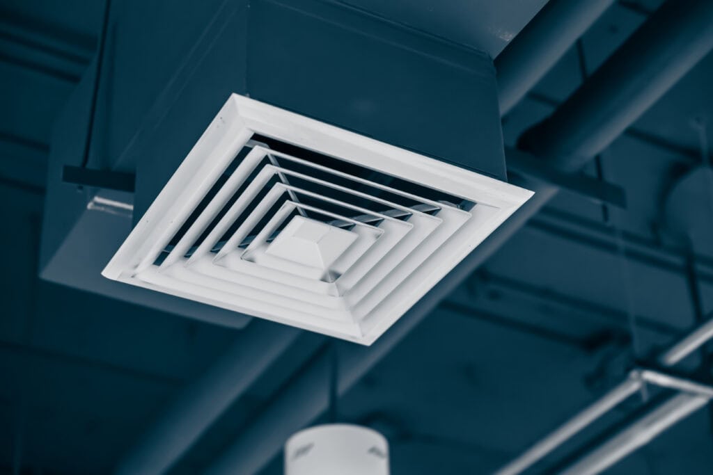 Ceiling-mounted air conditioning vent with a square design, set against a dark industrial ceiling background, where visible pipes and lighting fixtures add to the ambiance. This sleek setup complements spaces aware of details like dryer vent cleaning for optimal airflow efficiency.