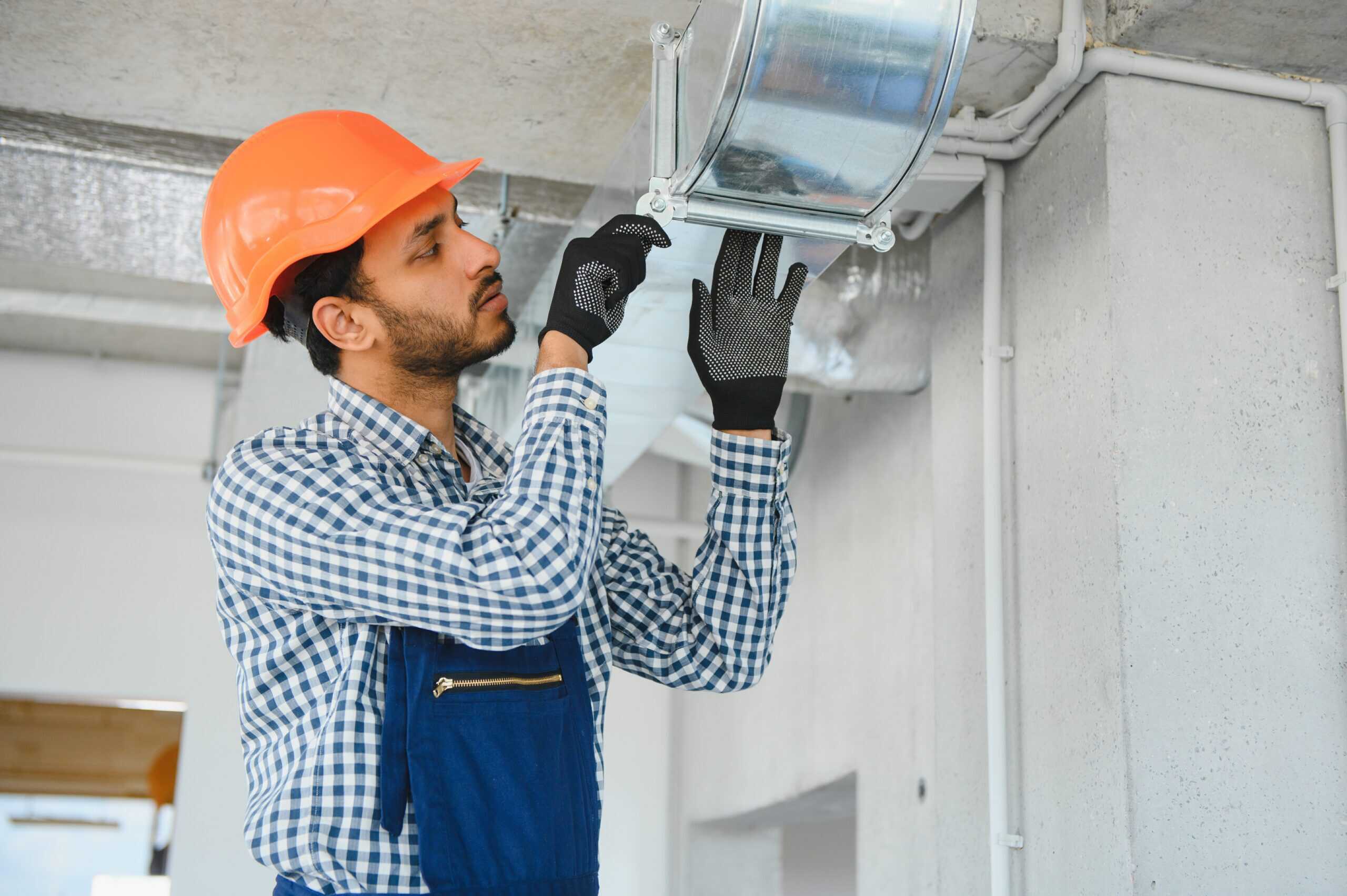 A construction worker wearing an orange hard hat and blue overalls is installing an air duct in a building. With his gloved hands, he adjusts the ductwork, ensuring everything is in place for seamless airflow, much like the precision required in gutter cleaning tasks.