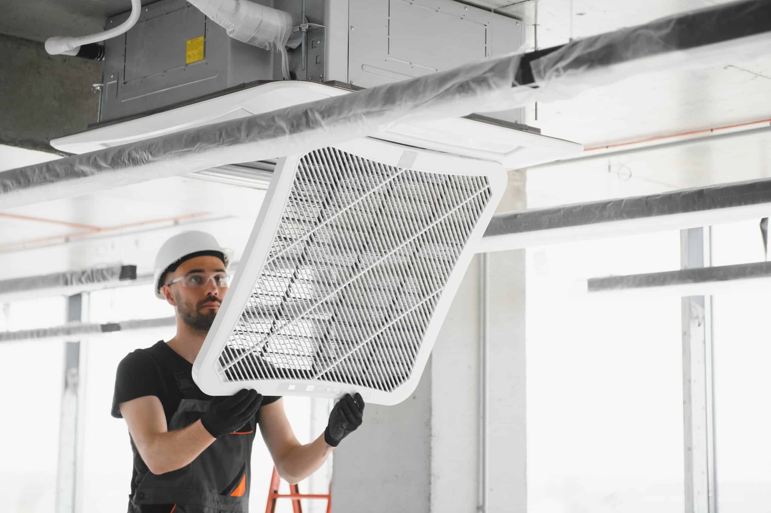 A construction worker wearing a hard hat and safety goggles is installing a white ventilation grille on a ceiling duct, possibly as part of dryer vent cleaning. He is on a ladder, working in a bright, modern interior space with large windows.