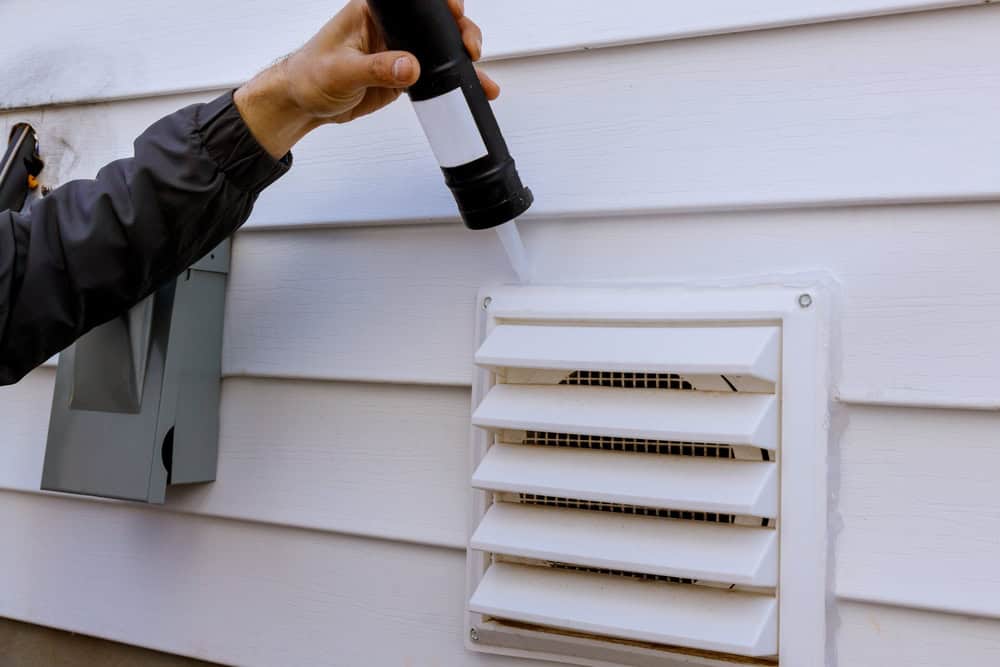 A person applies white caulk around a white exhaust vent on the side of a house using a caulking gun. The house exterior is covered in white horizontal siding, and an electrical box is visible in the background.