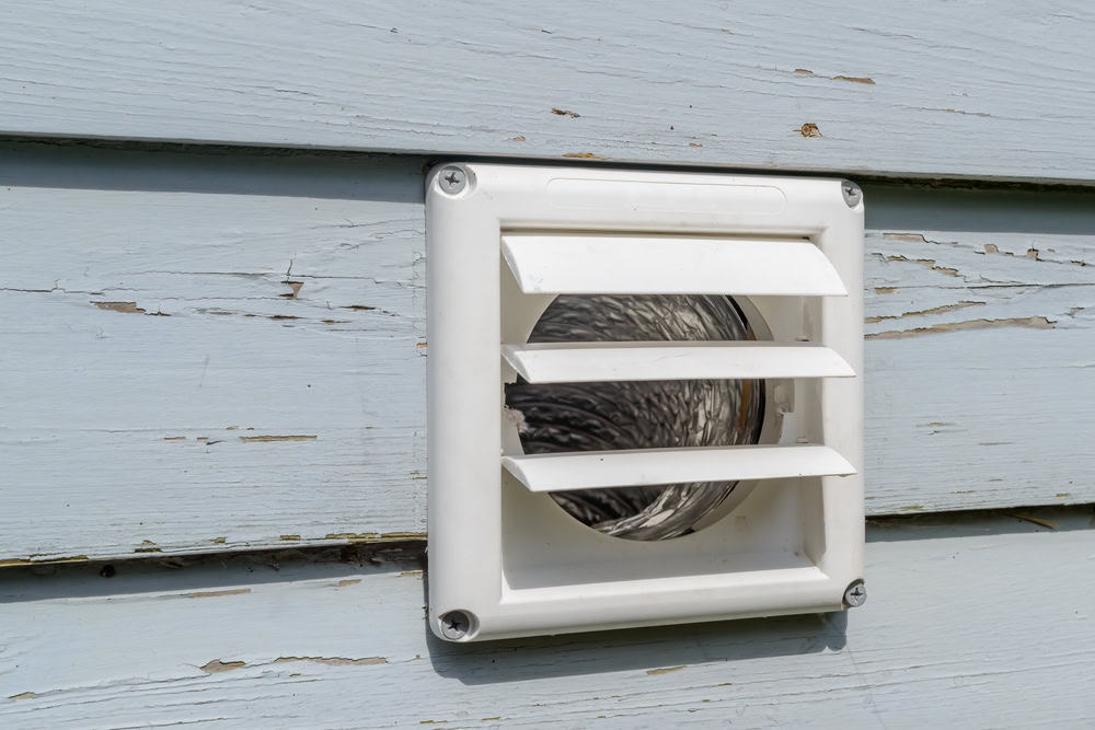 A white plastic dryer vent with slats is installed on a wooden exterior wall with peeling light blue paint.