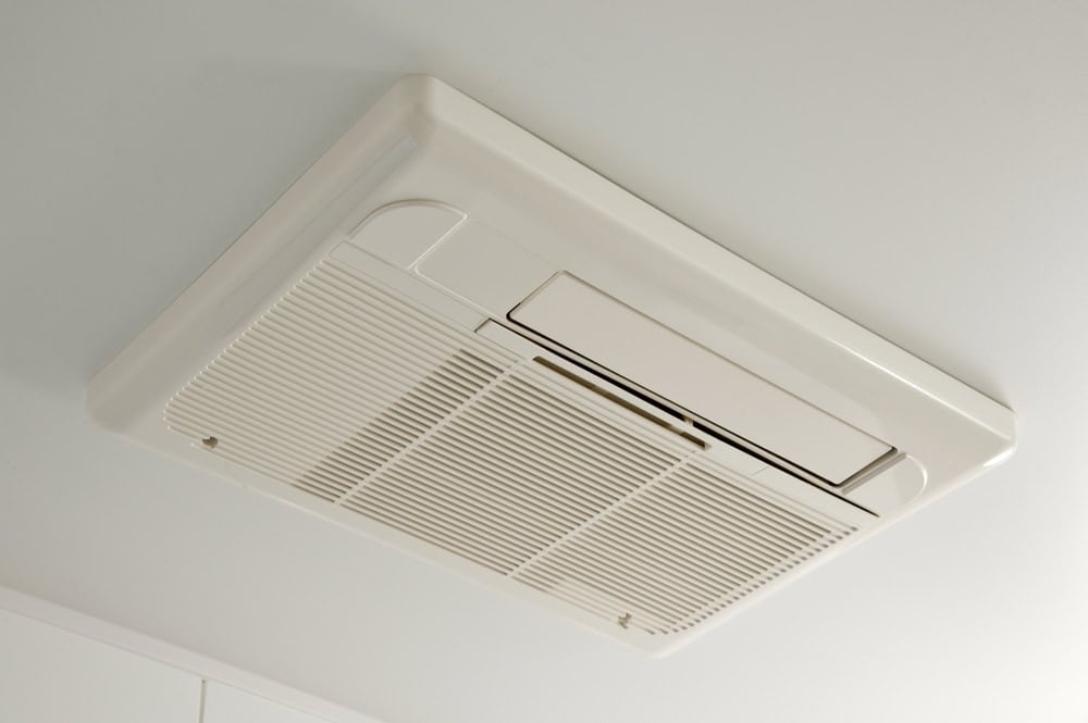 A white, rectangular ceiling-mounted ventilation unit with slatted grille and a small panel on one side. The unit is installed flush with the ceiling surface.