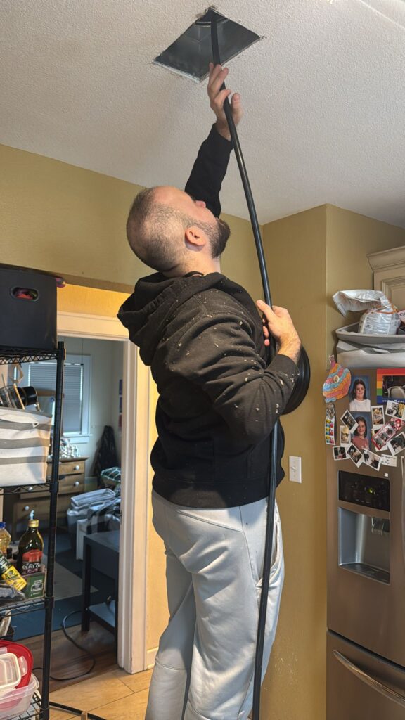 A person standing on a stool in a kitchen uses a long black cable to access a ceiling panel. The room has beige walls, a stainless steel refrigerator with magnets, and various items on the counter. The person wears a black hoodie and gray pants.