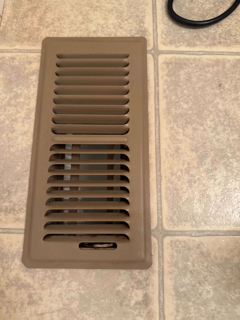 A rectangular beige metal floor vent with horizontal slats is set on a tiled floor with a beige and gray pattern. A black cable is partially visible in the upper right corner.