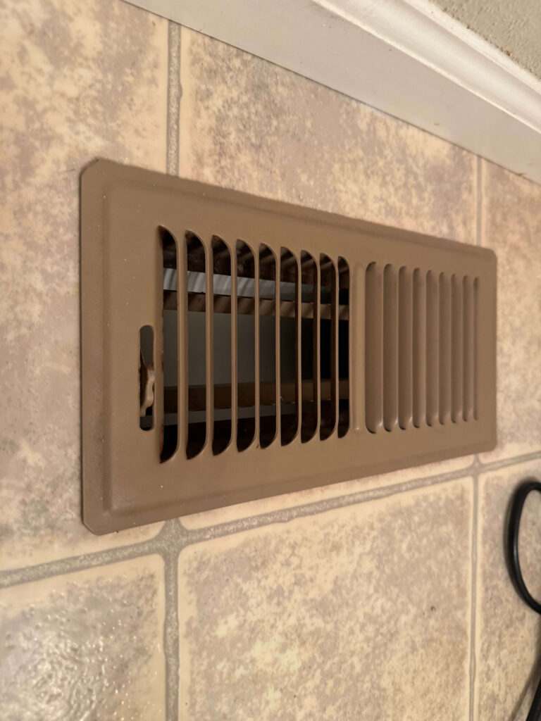 A beige floor vent is set into light-colored tile flooring. The vent cover has evenly spaced slats and is partially open, revealing the duct beneath. A small part of a black cable is visible in the lower right corner.