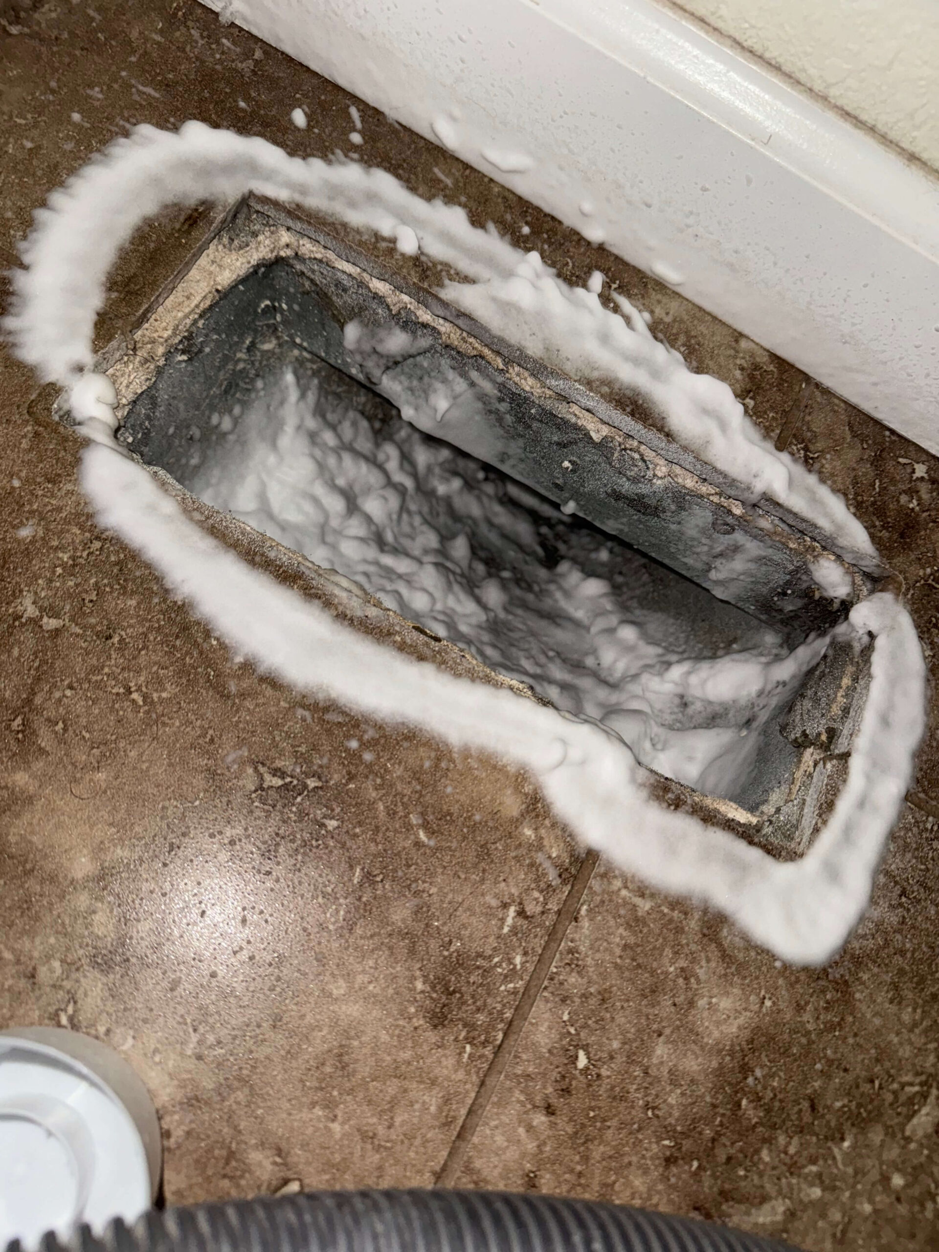 An open floor vent with dirt and dust build-up around the edges on a tiled floor. Cleaning foam is applied around the vent's perimeter, creating a thick white outline. A vacuum cleaner hose is partially visible in the corner.