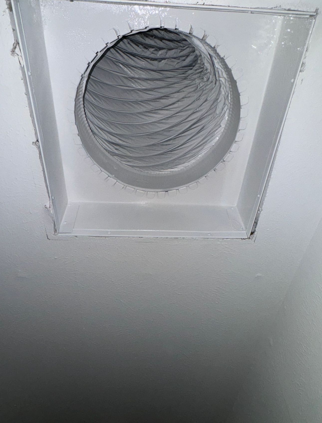 A white ceiling with a recessed square opening, housing a circular, flexible silver duct. The angular edges contrast with the spiraled texture of the duct inside the opening.