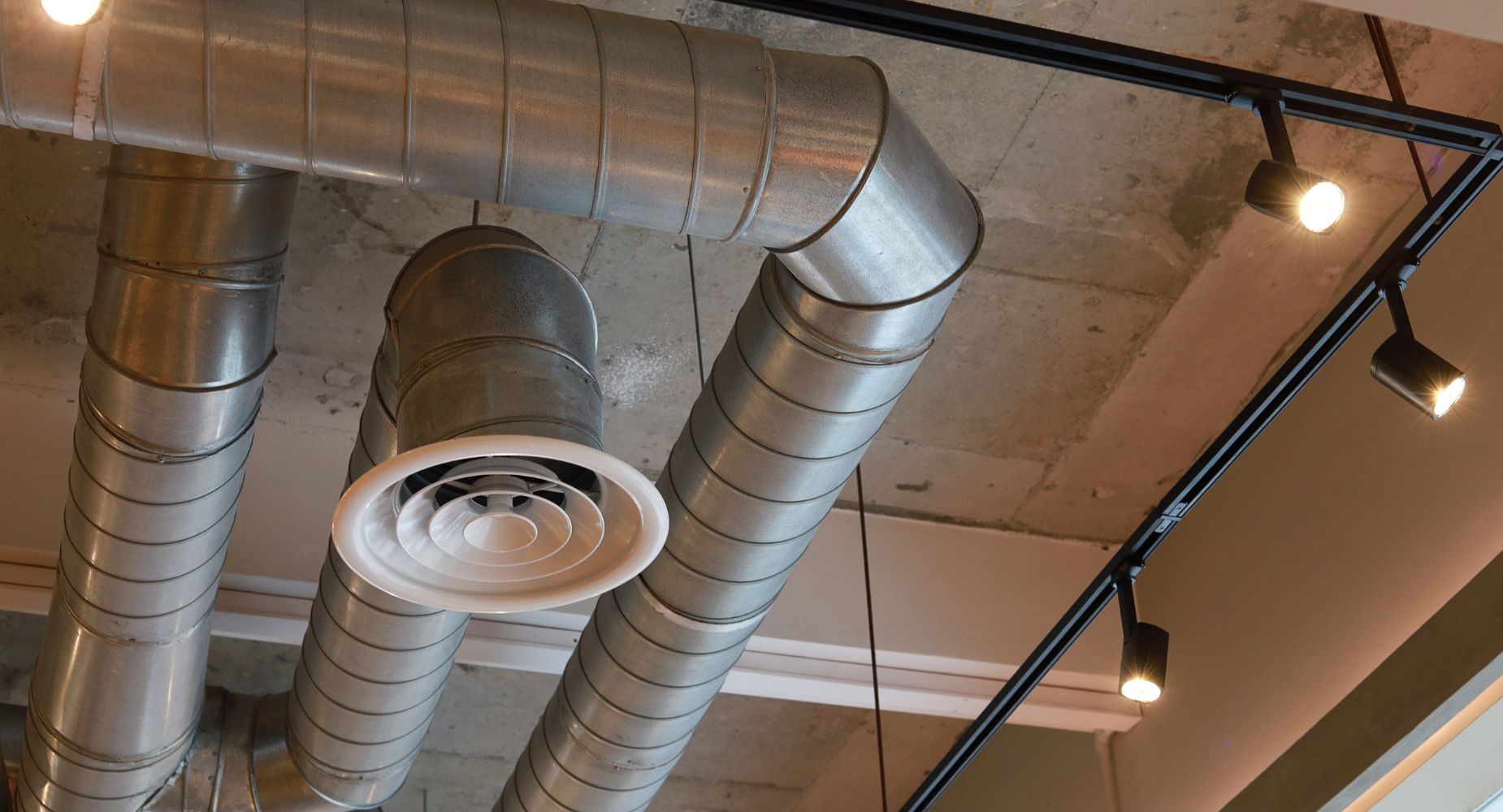 Exposed industrial ceiling with large metal air ducts and a circular vent. Track lighting with multiple spotlights illuminates the concrete ceiling and ducts.