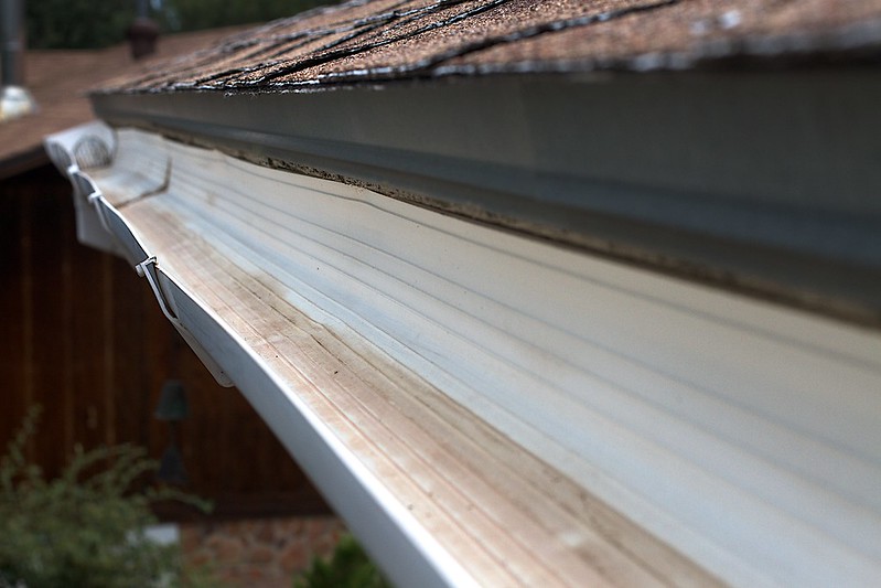 Close-up view of a roof gutter, showing streaks and dirt buildup inside. The gutter, reminiscent of neglected air ducts, has a slight bend and is attached to the roof's edge, with trees and a wooden building in the backdrop.