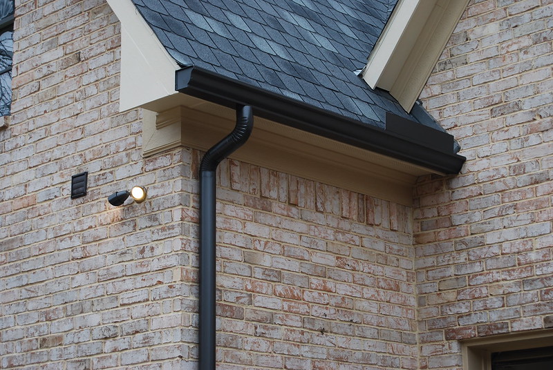 Close-up of a brick house corner with black roofing and a black downspout. A small outdoor light is mounted on the wall beside the drainage pipe, perfect for illuminating any late-night air duct cleaning projects in Denver, CO. Roof shingles are dark gray, and the bricks have a light beige tone.