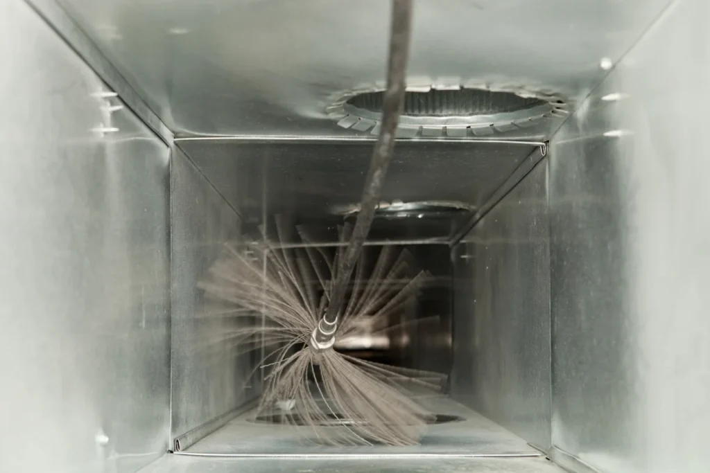 A spinning brush cleans the inside of a metal air duct, removing dust and debris as part of an HVAC maintenance process.