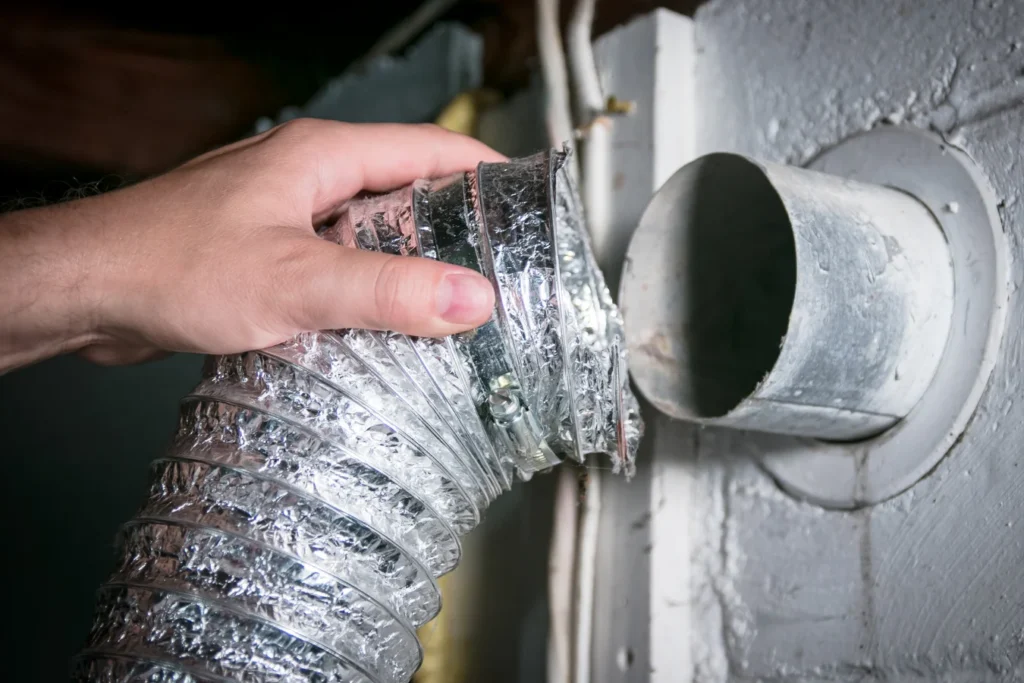 A hand is connecting or disconnecting a flexible silver dryer vent hose to a round metal wall duct opening.