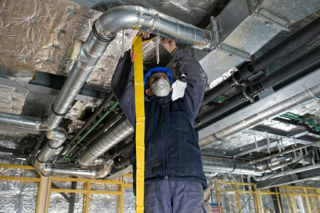 A construction worker wearing a blue helmet and face mask installs or inspects overhead metal ductwork and yellow insulation tape inside a building under construction, surrounded by exposed pipes and foil insulation.