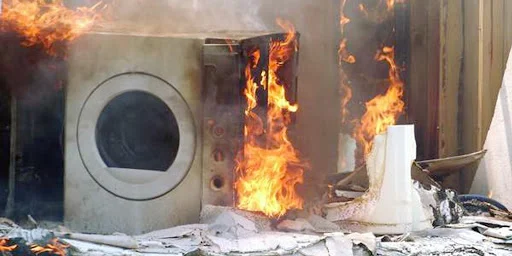 A washing machine on fire, surrounded by bright orange flames and smoke, with debris scattered around in front of a building or structure.